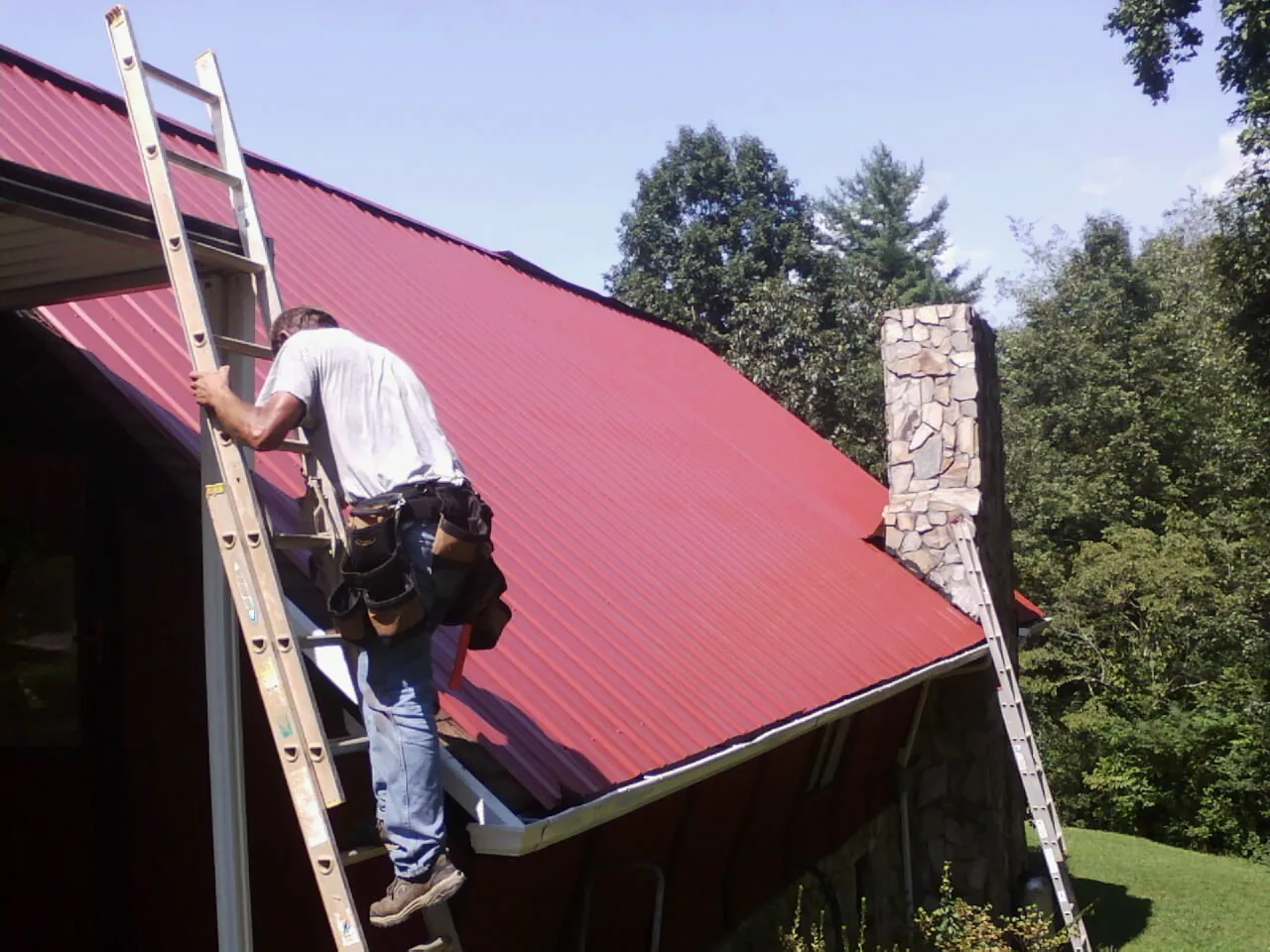 Roofer on a ladder beside a red metal roof and stone chimney — Soffit & Fascia Repair by Roofing Brothers in Huron